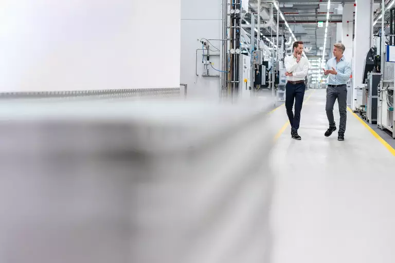 Symboldbild Sparringspartner – Zwei Männer unterhalten sich beim Gehen durch eine Produktionshalle